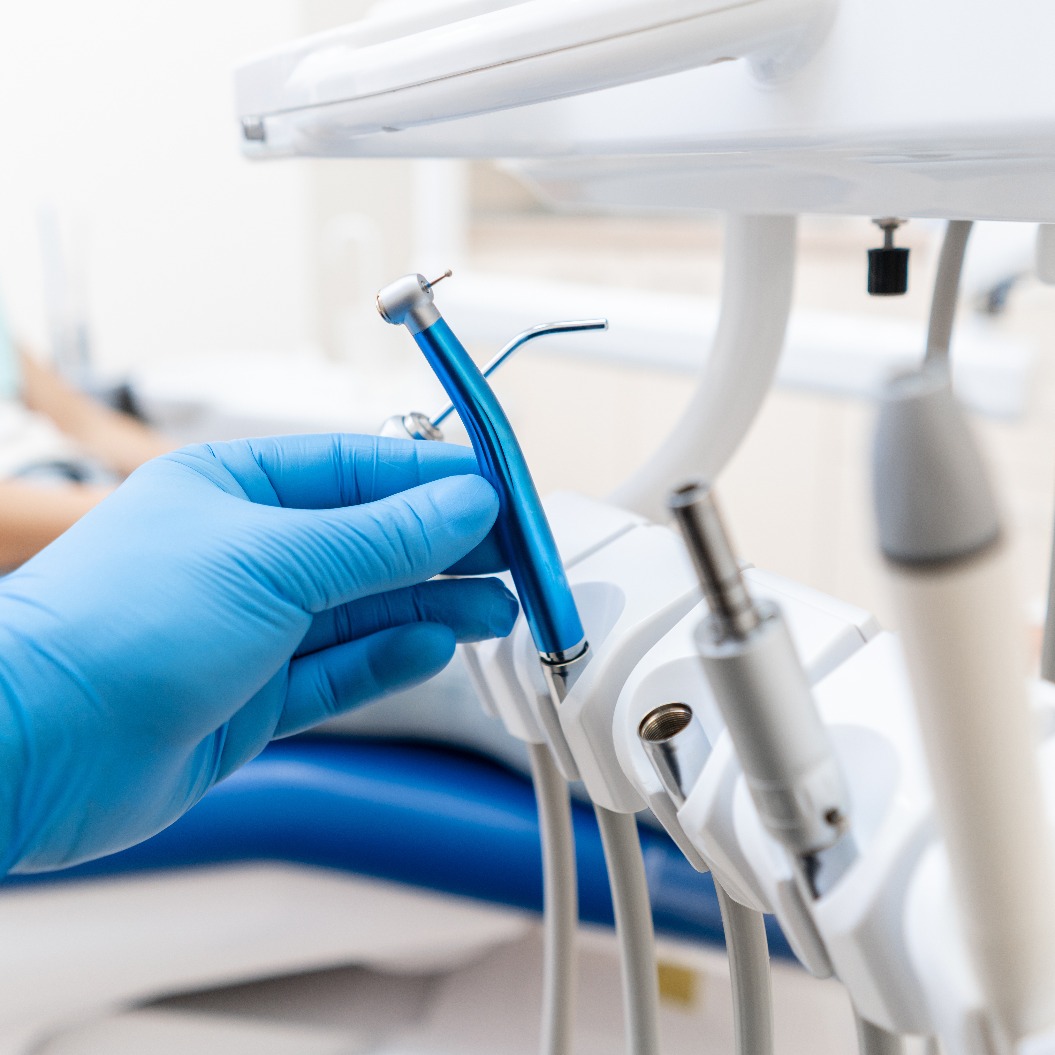 Nahaufnahme der Hand eines Zahnarztes im Handschuh, die eine zahnärztliche Hochgeschwindigkeitsturbine hält. Im Hintergrund ist der Patient auf dem blauen Stuhl zu sehen. Praxisraum, in dem der Zahnarzt die Untersuchung durchführt und seine Schlussfolgerungen zieht.