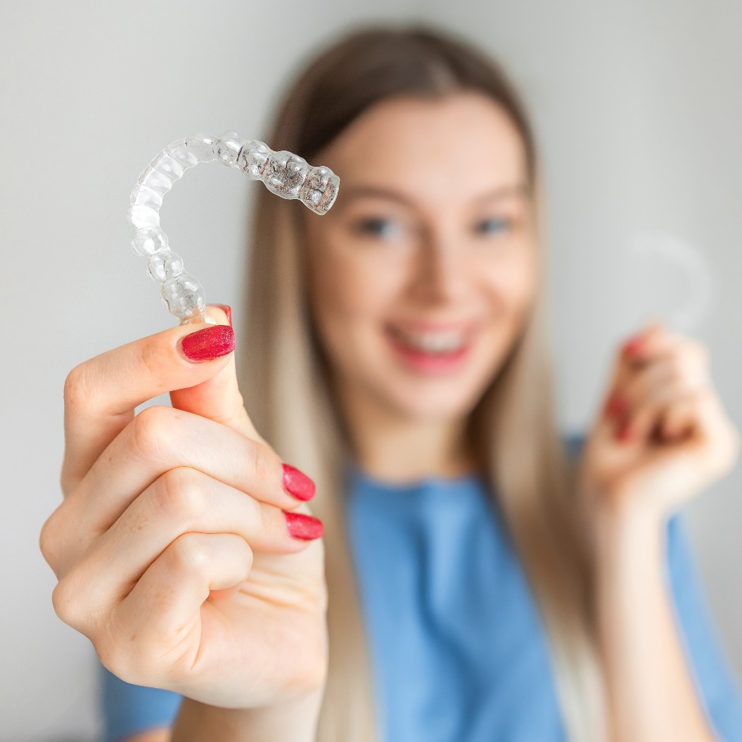 Eine weibliche Hand hält „Invisalign“, den unsichtbaren Aligner zur Zahnkorrektur, vor dem Hintergrund einer Frau im Profil. Konzept der Zahnkorrektur