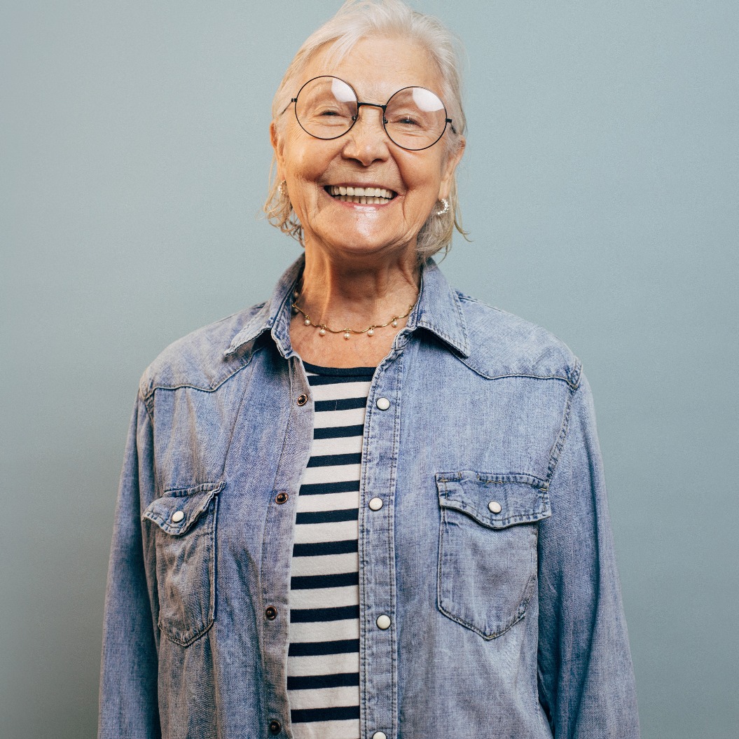 Fröhliche, sehr emotionale ältere Dame mit grauen Haaren, die eine Jeansjacke und ein Hemd trägt, in die Kamera blickt, lacht und vor einem blauen Hintergrund posiert. Elegante, glückliche Seniorin. Konzept: Glücksgefühle.