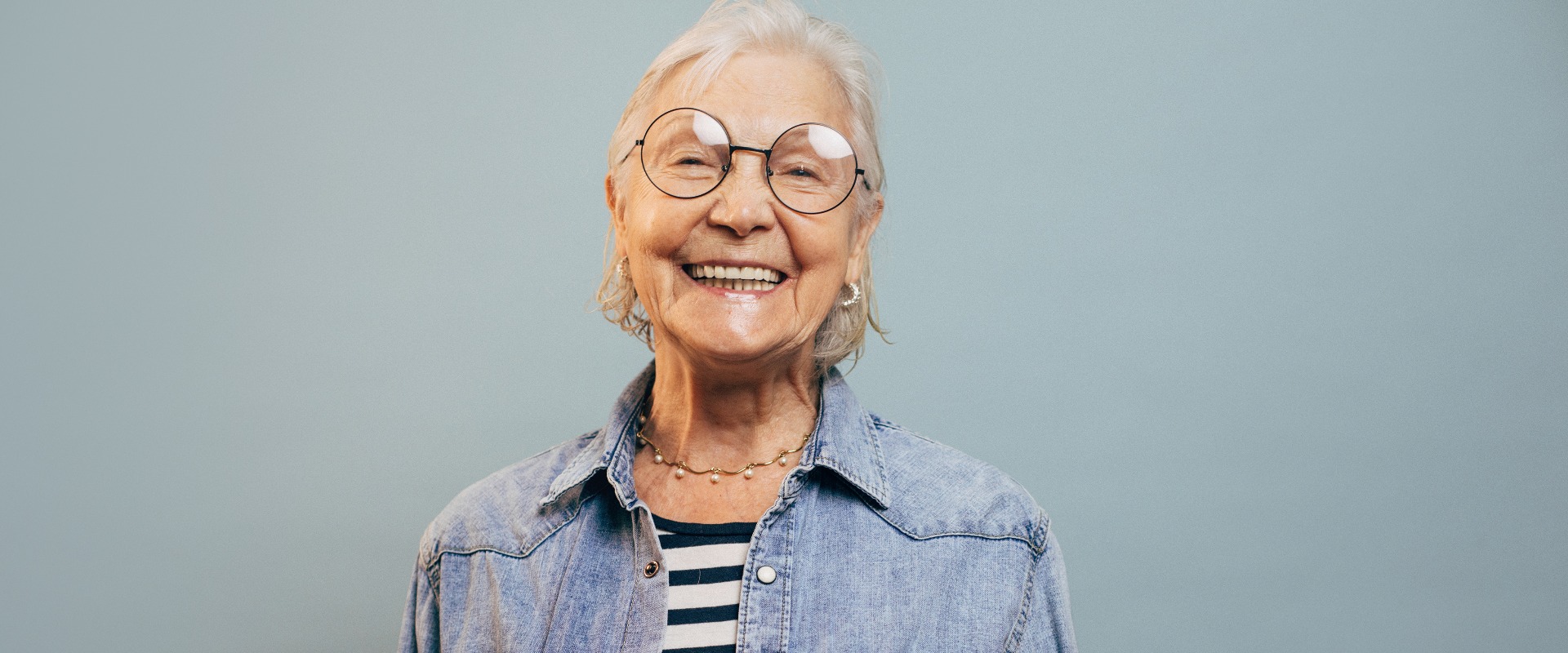 Fröhliche, sehr emotionale ältere Dame mit grauen Haaren, die eine Jeansjacke und ein Hemd trägt, in die Kamera blickt, lacht und vor einem blauen Hintergrund isoliert posiert. Elegante, glückliche ältere Dame. Konzept: Glücksgefühle.